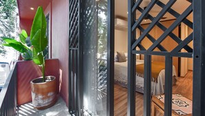 Un balcón con una planta en maceta, una puerta corrediza de vidrio que da a un dormitorio con una cama, y suelo de madera.