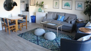 A modern living room with a grey sofa, wooden dining table, and chairs. There are two round ottomans on a patterned rug. - Steps to Boardwalk & Ocean! (Rehoboth Beach)