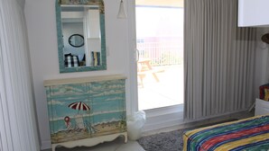 A bedroom with a beach-themed dresser, a colorful bedspread, a large window with curtains, and a view of the outside balcony. - Fantastic Views of Ocean from the Extra-large Patio! (Rehoboth Beach)