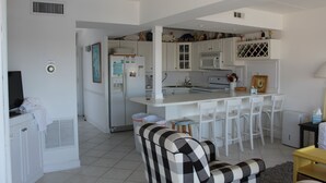 A compact kitchen with white cabinetry, a microwave, and a refrigerator. There are two bar stools and a small table. - Fantastic Views of Ocean from the Extra-large Patio! (Rehoboth Beach)