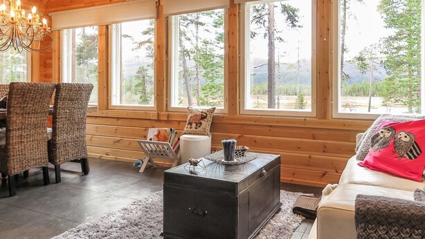 En mysig stuga med en matplats, en soffa, en öppen spis och stora fönster som ger utsikt över skogen.