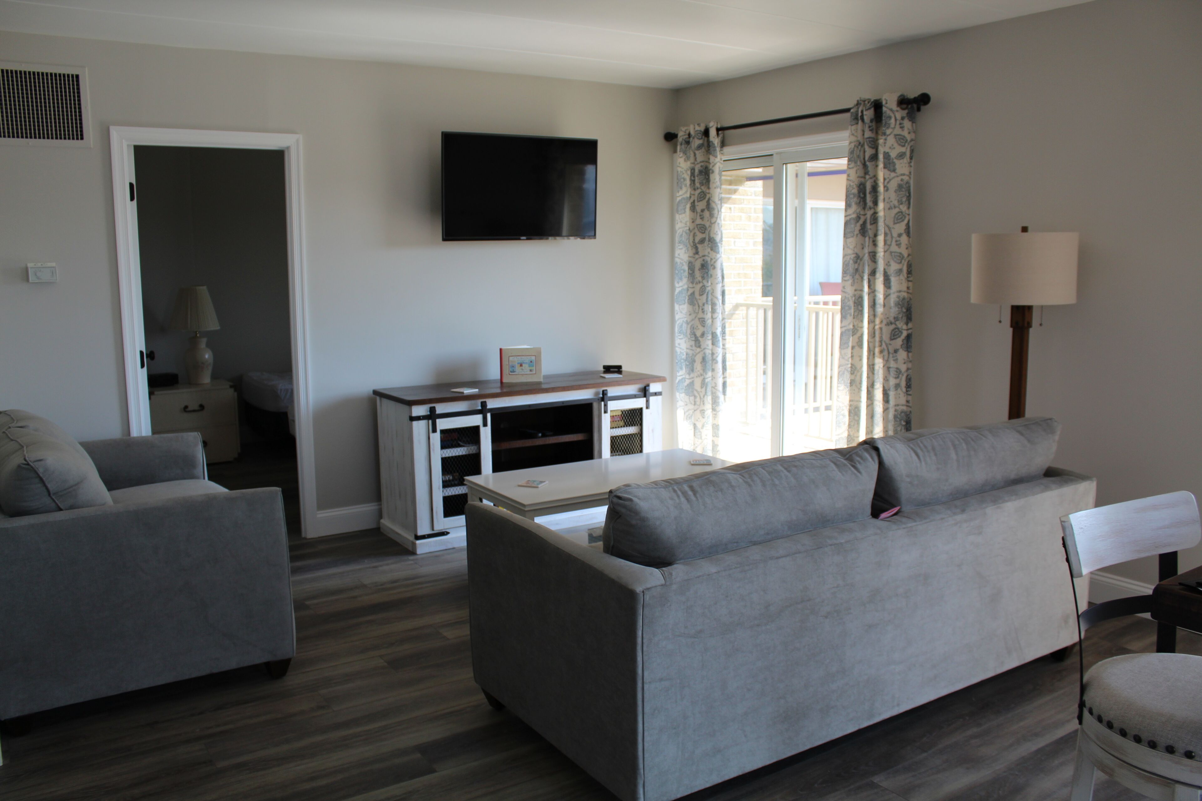 Un salón con un sofá gris, un televisor montado en la pared, una ventana con cortinas y una lámpara.