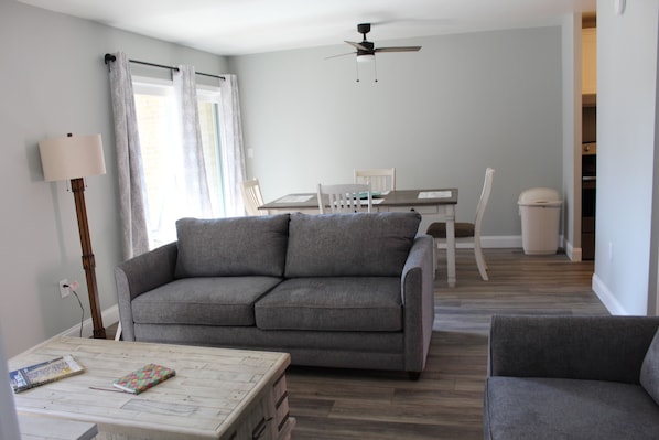 Un salon avec un canapé gris, une petite table et un coin repas comprenant une table et des chaises.