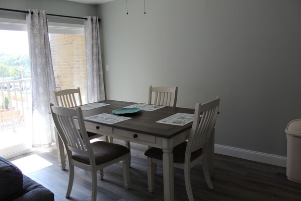 A dining area with a table set for four, chairs, a window with curtains, and a view of a brick building. - Charming Condo! Steps to Boardwalk! (Rehoboth Beach)