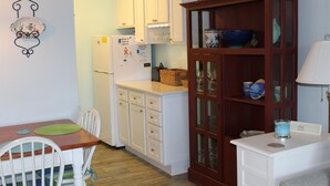 A compact kitchen with white cabinets, a wooden dining table with chairs, and a cabinet with glass doors containing bowls and glasses. - Beautiful Condo! Ocean Block & View! (Rehoboth Beach)