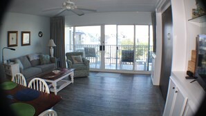 A spacious living area with a ceiling fan, a sofa, a dining table with chairs, and a balcony with a view of houses. - Beautiful Condo! Ocean Block & View! (Rehoboth Beach)