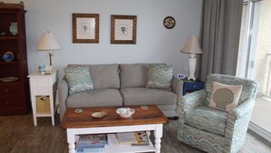 A living room with a sofa, a chair, a coffee table, a lamp, and a bookshelf. - Beautiful Condo! Ocean Block & View! (Rehoboth Beach)