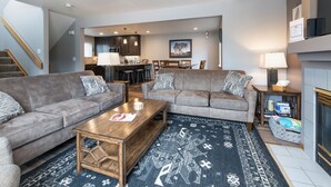 A modern living room with two grey sofas, a wooden coffee table, and a patterned rug. A kitchen is visible in the background with dark cabinets and a dining area. - Blue Granite Off Main Townhome: Updated, Great Location (Frisco)