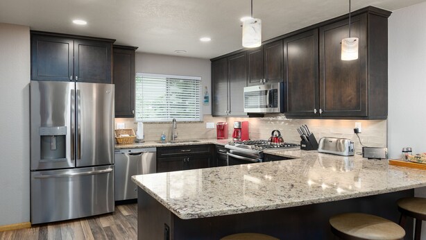 A modern kitchen with dark cabinets, stainless steel appliances, a granite countertop, and bar stools. - Blue Granite Off Main Townhome: Updated, Great Location (Frisco)