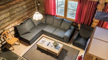 Un intérieur de chalet chaleureux avec un canapé d’angle, une table basse en bois, un fauteuil et une fenêtre avec des rideaux.