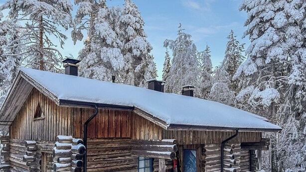 Une cabane en rondins, dont le toit est recouvert de neige, avec une cheminée, entourée d’arbres chargés de neige.