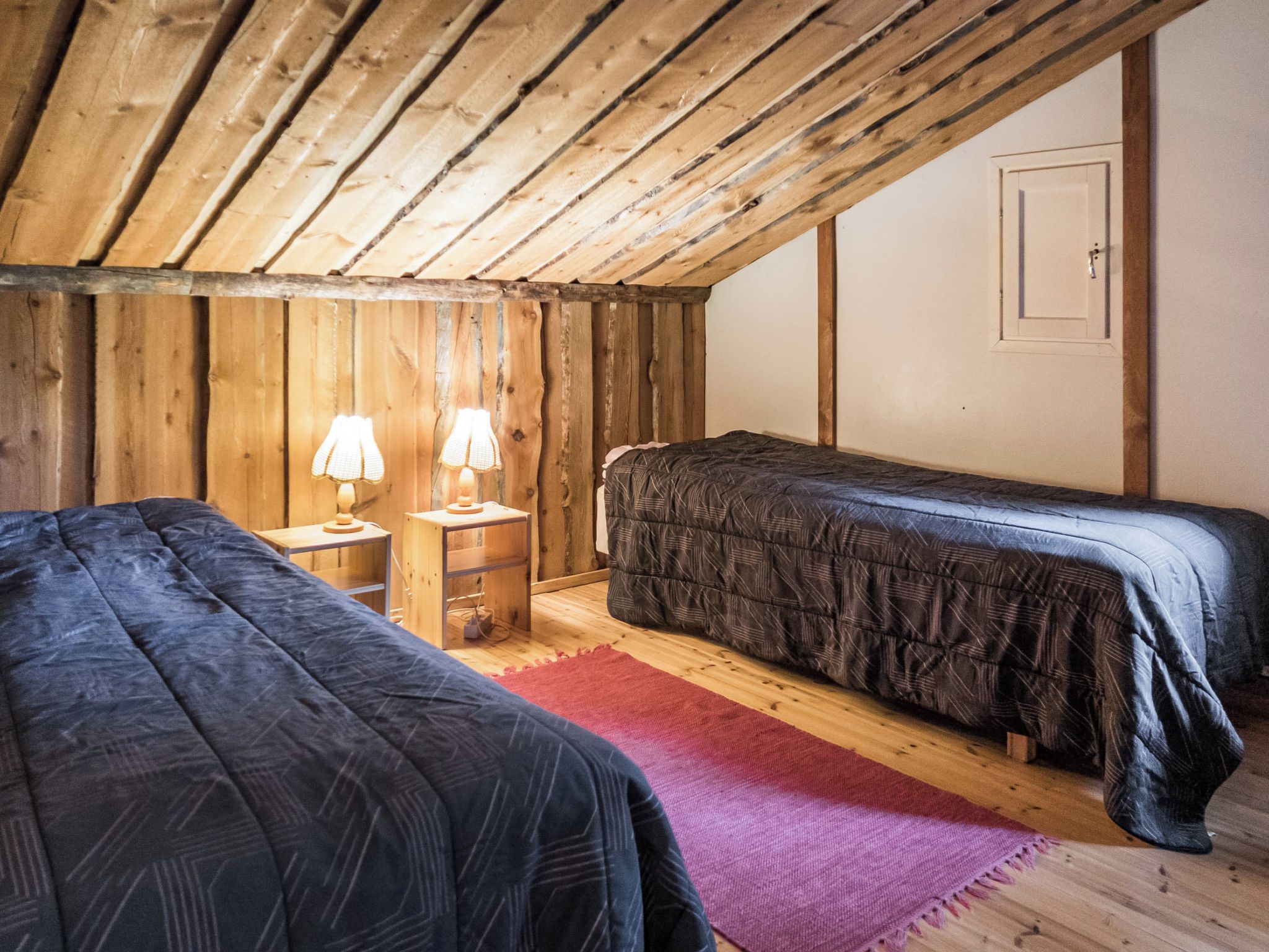 Une petite pièce chaleureuse aux murs en bois et au plafond incliné. On y trouve deux lits avec une literie sombre, une petite table de chevet en bois avec des lampes, et un tapis rouge sur le sol.