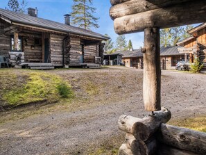 Une cabane rustique en rondins de bois, dotée d’un porche, d’une allée en gravier et bordée par une forêt.