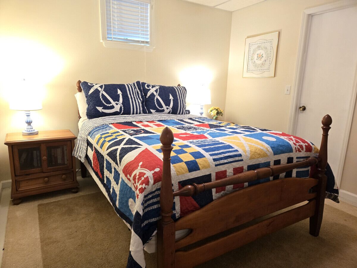 Une chambre à coucher avec un lit en bois, une table de chevet, une lampe, une fenêtre avec des stores et un tableau encadré au mur.