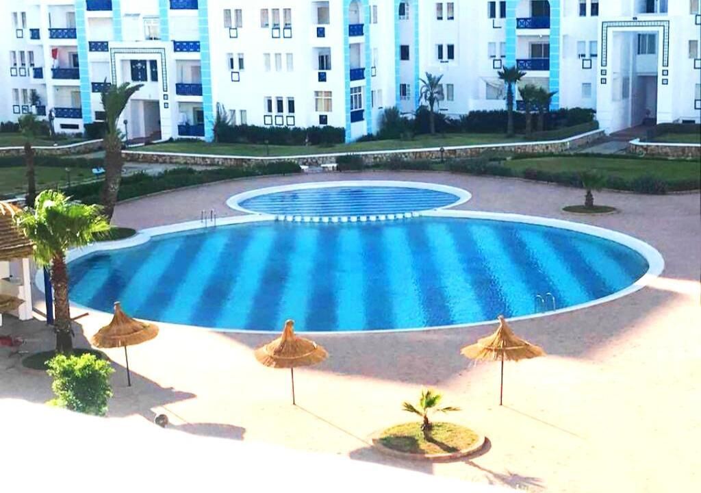 A swimming pool with a circular design, surrounded by palm trees and thatched umbrellas.