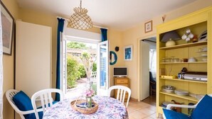 A dining area with a table set for four, surrounded by white chairs with blue cushions. A decorative basket with flowers is on the table. - 1-ROOM APARTMENT "La Creperie" (lege cap ferret)