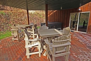 Un patio exterior con sillas Adirondack de madera y una mesa, una parrilla de barbacoa, y una puerta de vidrio que da a una zona cubierta.