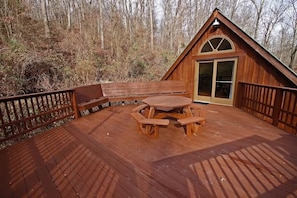 Una terraza de madera con una mesa de picnic y bancos, rodeada por una valla de madera, y una cabaña al fondo.