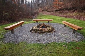 A circular fire pit surrounded by three wooden benches, set in a gravel area with grass and trees in the background. - Hidden Valley - modern, rustic, unique (Rockbridge)