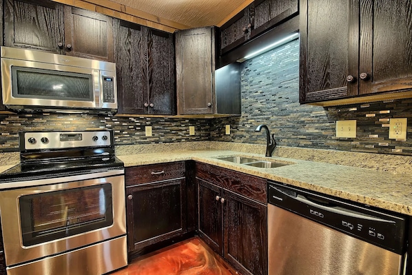 Una cocina con gabinetes de madera oscura, un microondas, horno y lavavajillas de acero inoxidable, y una encimera de granito.