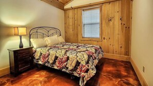 Un dormitorio con techo de madera, una cama con edredón floral, una mesita de noche con lámpara y una ventana con persianas.