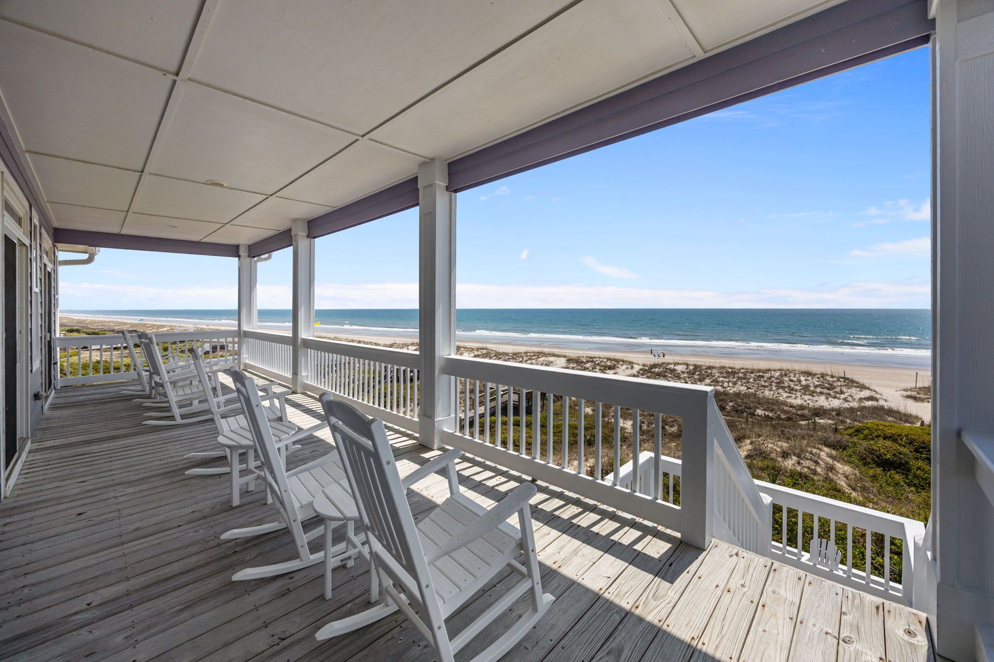 A covered porch with white rocking chairs and a view of the beach.