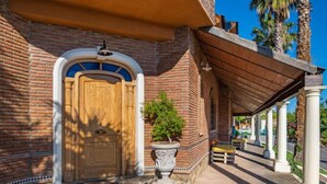 A brick building with a wooden door, a stone planter with a tree, and a covered walkway with columns. - El Balcon by Interhome (Córdoba/Baena)