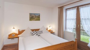 Un lit en bois avec une literie blanche, deux tables de chevet avec des lampes, une table de nuit, une fenêtre avec des rideaux et un tableau accroché au mur.