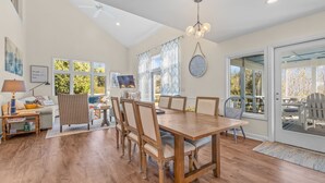 A spacious dining area with a wooden table and chairs, a ceiling fan, and a large window offering a view of trees.