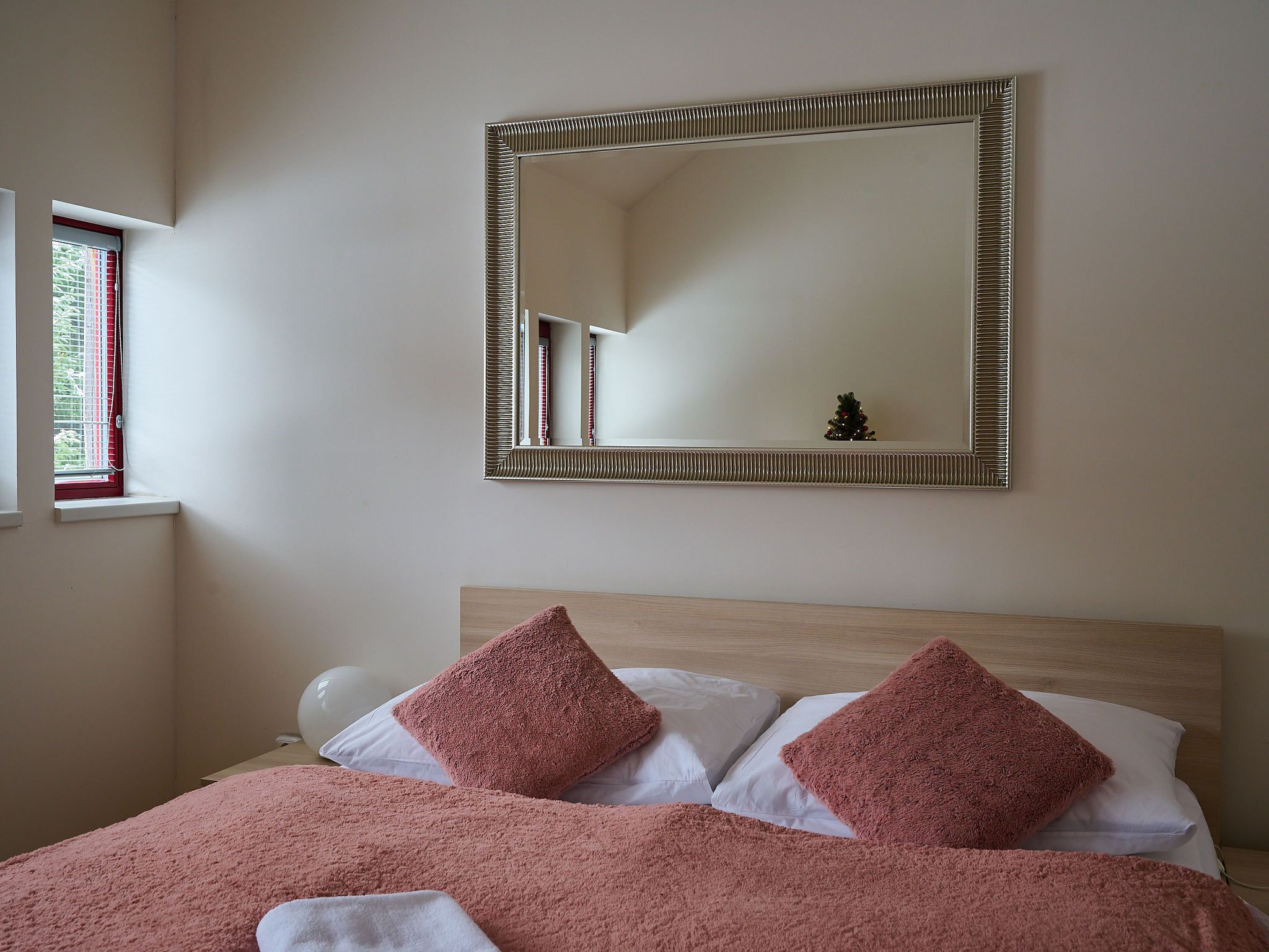 A bedroom with a bed, two pink pillows, a white blanket, a wooden headboard, a mirror with a decorative frame, and a window with blinds.