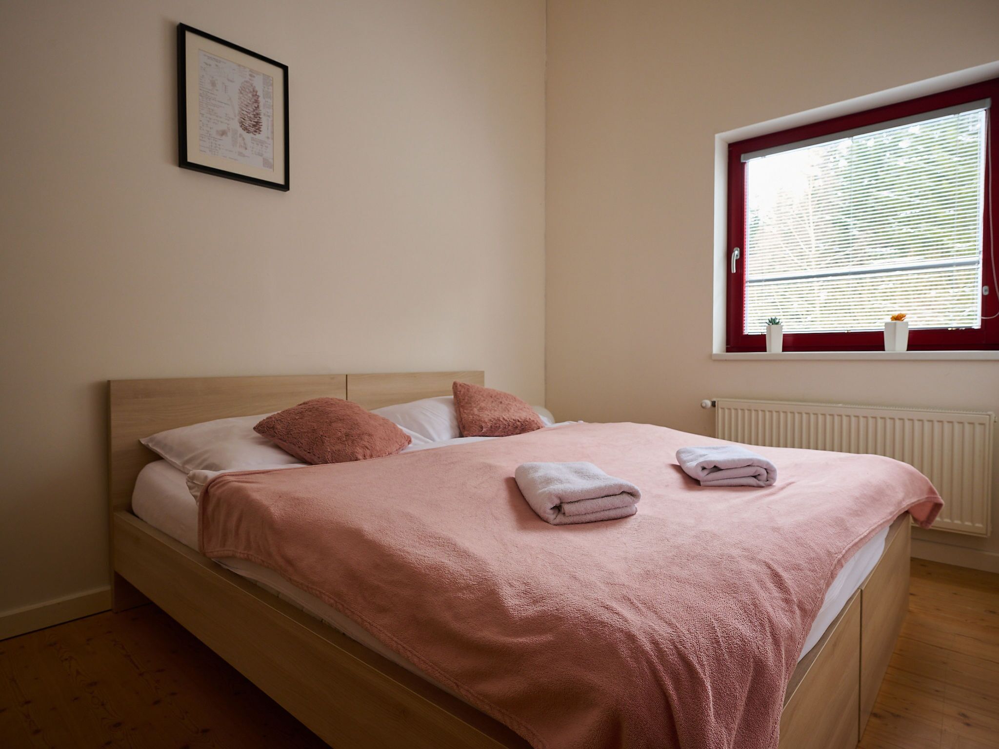 Un letto singolo con una coperta rosa, due cuscini e due asciugamani piegati. C'è un quadro appeso al muro e una finestra con persiane.