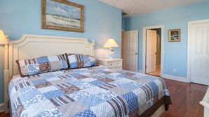 A bedroom with a large bed, a nightstand, a lamp, a door, and a framed picture on the wall.
