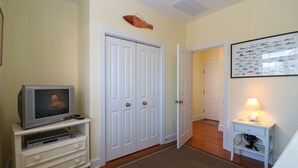 A room with a television on a white stand, a closed white door, a framed fish picture on the wall, and a small table with a lamp.