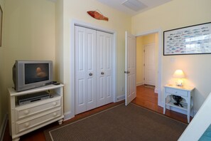 A room with a television on a white stand, a closed white door, a framed fish picture on the wall, and a small table with a lamp.