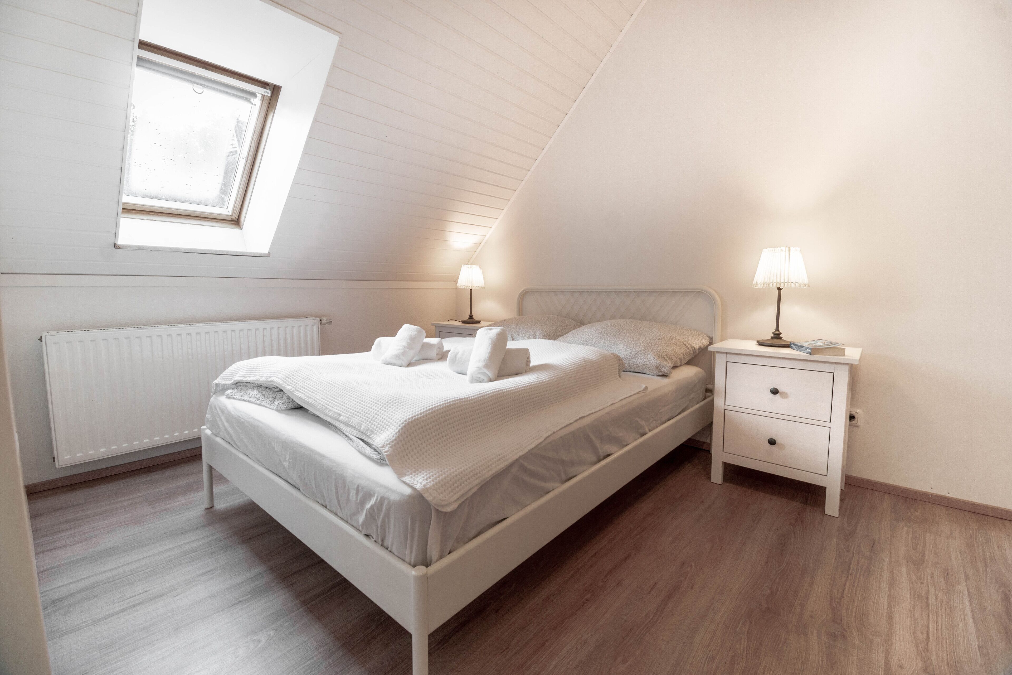 Un dormitorio con techo inclinado, una cama con ropa blanca, dos mesitas de noche con lámparas y un radiador.