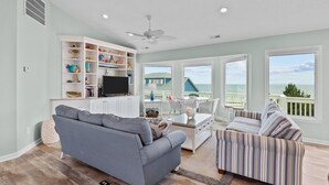 Ein Wohnzimmer mit grauem Sofa, gestreiftem Sofa, Fernseher und Bücherregal. Große Fenster bieten Blick aufs Meer.