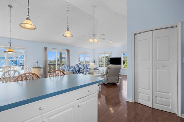 A spacious kitchen with a blue countertop, white cabinets, and hanging pendant lights. There is a dining area with a table and chairs, and a TV in the background.