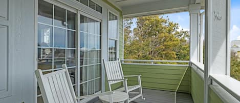 Eine überdachte Veranda mit zwei weißen Adirondack-Stühlen und Blick auf Bäume und Gebäude im Hintergrund.
