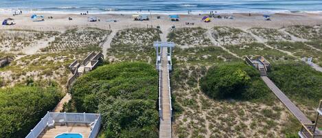 A beach with a wooden boardwalk leading to a pool area, surrounded by dunes and beach umbrellas.