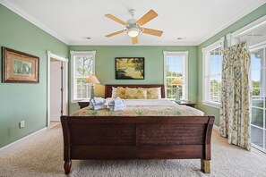 Une chambre à coucher avec un lit, un ventilateur de plafond, deux lampes fixées au mur, un tableau encadré et une fenêtre avec des rideaux.