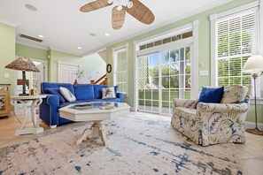 A living room with a blue sofa, a patterned armchair, a glass coffee table, and a ceiling fan. - Oceanside Interior Home,Private Pool,Golf Cart,Beach Access,in Spinnaker's Reach (Emerald Isle)