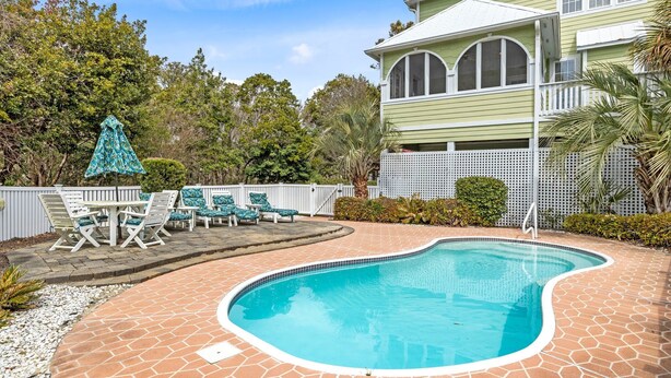 Une piscine avec une terrasse carrelée, entourée d’une clôture blanche, et un espace patio aménagé avec des chaises et une table.