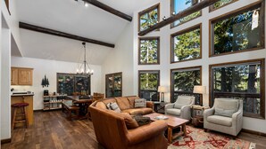 A spacious living area with a brown leather sofa, a wooden coffee table, and a patterned rug. There are multiple windows providing views of the outdoors. - Spacious Whitefish Mountain residence with some of the best ski in/out on the mountain. (Whitefish)