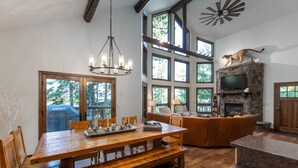 A spacious living area with a wooden dining table, a stone fireplace, and a large window overlooking a forest. - Spacious Whitefish Mountain residence with some of the best ski in/out on the mountain. (Whitefish)