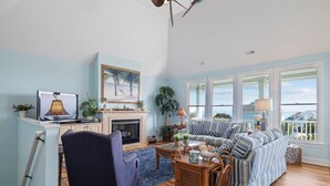 A living room with a fireplace, a sofa set, a dining table, and a view of the ocean through large windows. - Oceanview with elevator, private pool, golf cart, in Spinnakers Reach community (Emerald Isle)