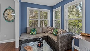 A room with a blue wall, a large window with blinds, a sofa with cushions, a wicker chair, and a table with a lantern. - Pet Friendly, Oceanside Interior Home Private Pool, Golf Cart, and Easy Access. (Emerald Isle)
