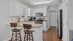 A kitchen with white cabinets, a granite countertop, two wooden bar stools, a stainless steel refrigerator, and a pendant light. - Pet Friendly, Oceanside Interior Home Private Pool, Golf Cart, and Easy Access. (Emerald Isle)