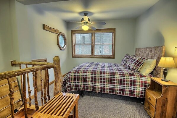 A bedroom with a bed, a wooden chair, a nightstand with a lamp, a ceiling fan, and a window with blinds.