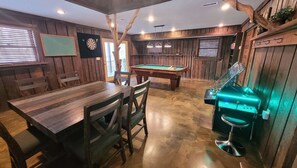 A wooden-paneled room with a pool table, a dartboard, and a bar area with a grill.