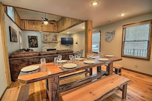 Un coin repas avec une table en bois rustique dressée pour six personnes, une cheminée en pierre et une grande fenêtre équipée de stores.